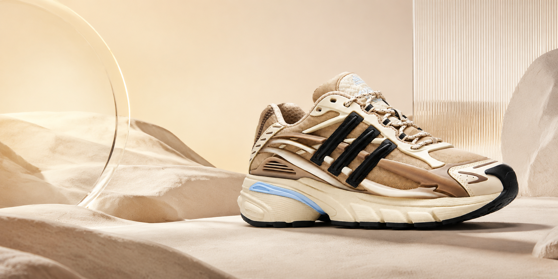 A beige and brown Adidas Yeezy Max sneaker with black three-stripes and a light blue midsole accent, displayed on a draped beige fabric surface with a glass vessel in the background.